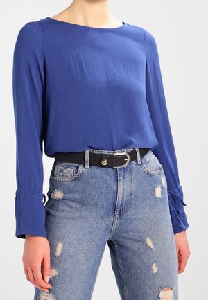 Vrouw draagt een lange mouw blauwe blouse, ingestopt in hooggetailleerde, gescheurde blauwe jeans met een zwarte riem met een zilveren gesp.