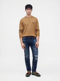 Jeune homme debout, les mains détendues, portant un pull marron, un jean bleu déchiré et des baskets vertes avec des lacets orange et bordeaux.