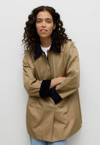 Chaqueta beige claro con cuello y puños azul marino, con cierre de botones y dos bolsillos delanteros, llevada sobre una camisa blanca.