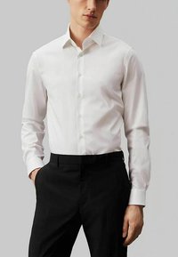 Camicetta bianca in tessuto liscio, dotata di colletto, bottoni frontali e maniche lunghe. Abbinata a pantaloni neri su misura.