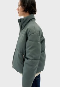 Kurtka puchowa w stonowanej zieleni, z wysokim kołnierzem, zamkiem błyskawicznym z przodu oraz dwoma bocznymi kieszeniami. Miękka, gładka tekstura z pikowanym wzorem.