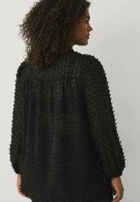 Zwarte, gestructureerde blouse met lange mouwen, voorzien van een gespikkeld patroon en gerimpelde details aan de achterkant. Lichtgewicht, vloeiende stof.