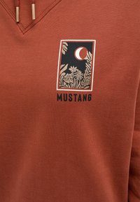 Sudadera de color óxido con un gráfico de plantas y una luna, junto al texto "MUSTANG" en negro. Textura suave con un diseño de cuello en V.