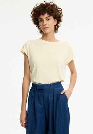 Mujer con cabello rizado que lleva grandes pendientes de aro, camiseta de manga corta color crema y pantalones azules de pierna ancha y talle alto, con una mano en el bolsillo.