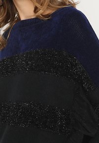 Musta ja laivastonsininen neule, jossa raidallinen kuvio; pehmeää, teksturoitua mustaa lankaa ja sileää laivastonsinistä kangasta. Pyöreä pääntie ja rento istuvuus.