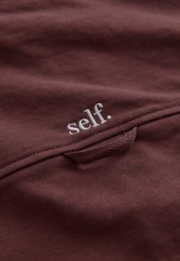 Brown fabric with white embroidered word "self." above a stitched fabric loop detail.