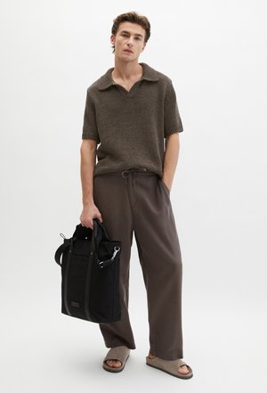 Man met een bruin gebreid shirt met korte mouwen, losse bruine broek, beige sandalen, draagt een zwarte tas, staand op een witte achtergrond.