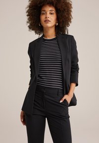 Žena s kudrnatými vlasy, která má na sobě zlaté náušnice ve tvaru kruhu, černý blazer, černobílý pruhovaný top a černé kalhoty, stojící se rukou v kapse.