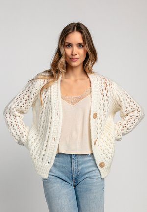 Gruby, biały kardigan z dzianiny o otwartym splotcie, z dużymi drewnianymi guzikami i prążkowanymi mankietami. Noszony na jasnym podkoszulku i dżinsach.