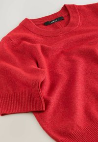 Red knitted sweater with short sleeves, featuring a ribbed round neck and hem. Soft texture with subtle variations in color. Brand label visible.