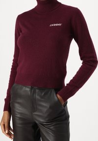 Kobieta w burgundowym swetrze z golfem z białym napisem "2ND DAY" i czarnymi skórzanymi spodniami, z jedną ręką w kieszeni, na neutralnym tle.