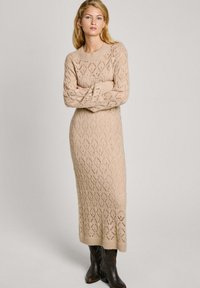 Abito in maglia beige con un motivo a diamante testurizzato, maniche lunghe e scollo a giro, che arriva fino alle caviglie, abbinato a stivali scuri fino al ginocchio.