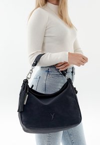Frau trägt ein weißes, geripptes Langarmoberteil und hellblaue Jeans, während sie eine große schwarze Handtasche mit einem Quastenanhänger und einem Metalletikett hält.