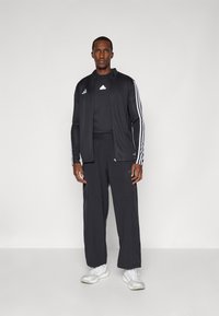 Survêtement de sport noir avec des rayures blanches sur les côtés, veste zippée et pantalon ample. Porté avec des baskets blanches.