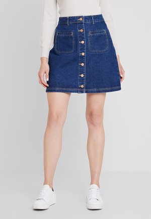 Vrouw die een blauwe denim rok met knopen aan de voorkant en twee voorzakken draagt, een witte top met lange mouwen en witte sneakers, staand tegen een effen achtergrond.