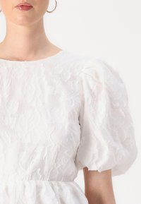 Λευκό φόρεμα με φουσκωτά μανίκια και λουλουδάτη κέντηση. Διαθέτει στρογγυλή λαιμόκοψη και υφασμάτινη υφή με συγκέντρωση στη μέση. Λείος τελειωμένος.