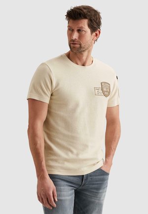 Moški v bež majici s kratkimi rokavi z obliži na prsih in modrih kavbojkah, stoji z eno roko v žepu, gleda levo.