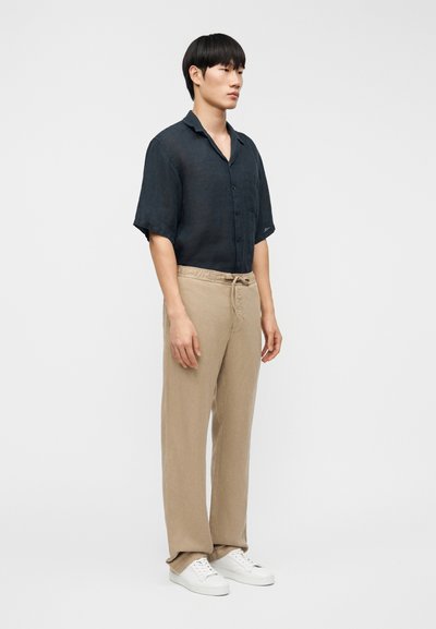 Νέος άνδρας όρθιος, φοράει μαύρο πουκάμισο με κοντά μανίκια και κουμπιά, μπεζ παντελόνι με κορδόνι και λευκά αθλητικά παπούτσια, σε απλό λευκό φόντο.