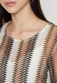Pleteni top z zigzag vzorom v krem, rjavi in črni barvi. Okrogla izreza, teksturirana tkanina in podrobnosti na ramenskem šivu so vidne.