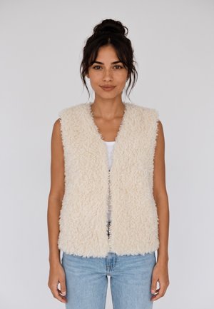 Donna con capelli scuri raccolti in uno chignon, che indossa un gilet soffice crema senza maniche sopra una maglietta bianca e jeans azzurri chiari su sfondo semplice.
