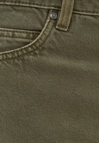 Tissu en denim vert olive avec une texture côtelée diagonale, des bords cousus et un bouton en métal gravé du logo de la marque.