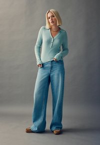 Haut bleu clair à manches longues avec un col et des détails de boutons, associé à un jean en délavé clair à coupe ample et des chaussures brunes à enfiler.