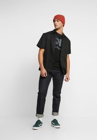 Black short-sleeve shirt with plaid pattern, worn over a black graphic t-shirt. Dark denim jeans, rolled cuffs, and green sneakers. Red beanie.