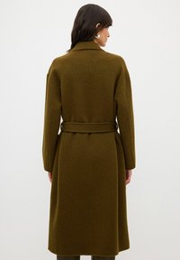 Olivgrön ullkappa med bälte i midjan, långa ärmar och rymlig silhuett, med klassisk krage och mjuk textur.