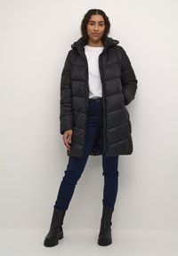 Manteau en duvet noir avec une capuche, présentant un design matelassé, une fermeture éclair à l'avant et deux poches sur les côtés. Associé à un jean bleu foncé et des bottes noires.