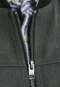 Gros plan d'une fermeture éclair d'une veste grise texturée partiellement ouverte, dévoilant une chemise blanche à rayures noires boutonnée en dessous.