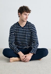 Marineblau-weiß gestreiftes Langarm-V-Ausschnitt-Oberteil, kombiniert mit passender marineblauer Hose, vor einem hellen Teppichhintergrund. Lässige Passform, weicher Stoff.