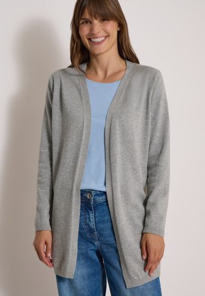 Femme souriante portant un cardigan gris clair ouvert devant, une chemise bleu clair et un jean bleu, debout devant un fond uni.