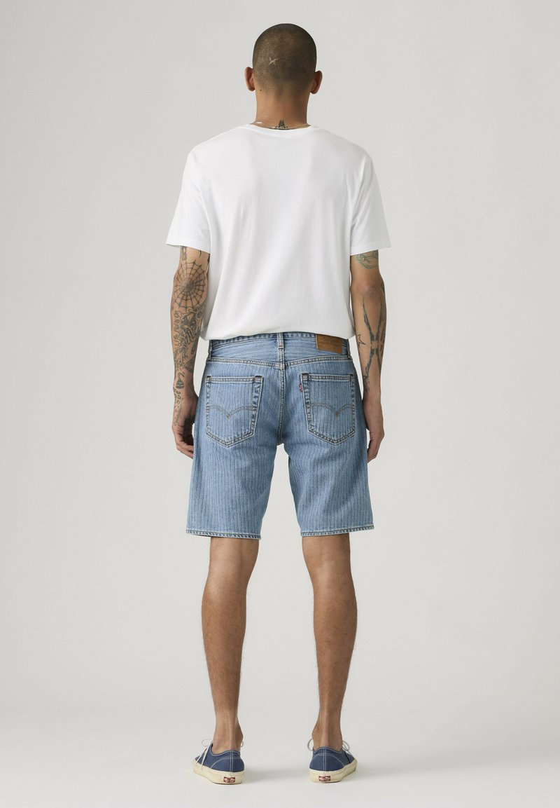 Hombre con los brazos tatuados, vistiendo una camiseta blanca, pantalones cortos de mezclilla azul claro y zapatillas azul marino, de pie de espaldas sobre un fondo liso.