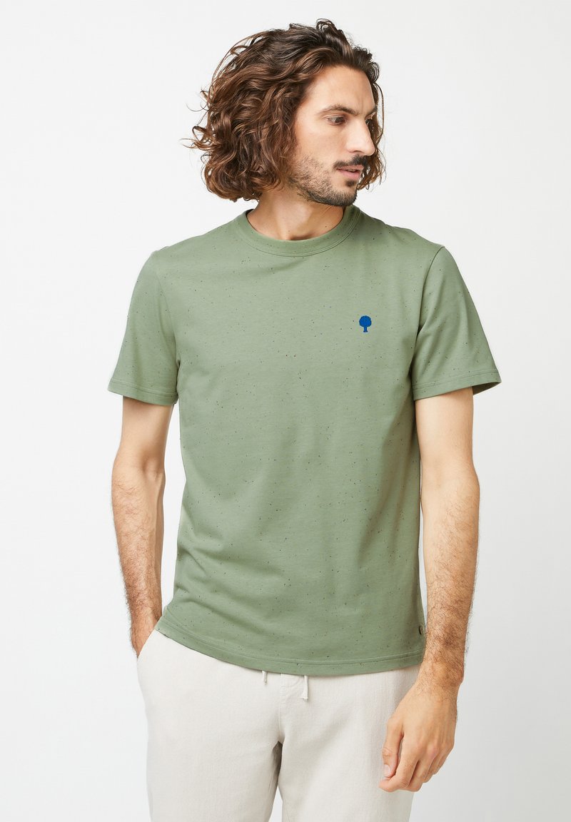 T-shirt en coton vert olive avec une texture mouchetée, col rond, manches courtes et un petit logo d'arbre bleu sur la poitrine.