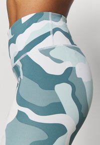 Leggings a vita alta con un motivo mimetico presentano tonalità di teal e bianco, realizzati in un tessuto elasticizzato e liscio con cuciture piatte per un comfort ottimale.