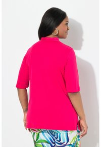 Heldroze shirt met korte mouwen en hoge hals, met een decoratieve rij studs bij de kraag. De stof lijkt glad en elastisch.
