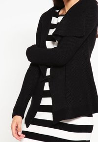 Svart stickad kofta med öppen front, texturerad tyg och bred krage, lager på en svart och vit randig klänning.