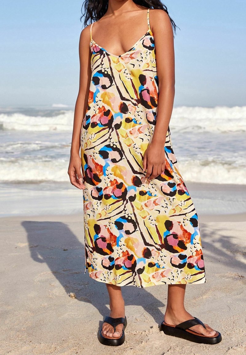 Robe maxi sans manches à motif floral dans des tons pastel de rose, jaune, bleu et noir. Portée avec des sandales plates noires sur une plage.