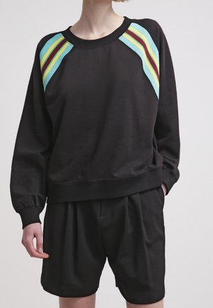 Person trägt einen schwarzen Sweatshirt mit blauen, gelben und roten Streifen auf den Schultern, kombiniert mit lockeren schwarzen Shorts, steht mit einer Hand in der Tasche.