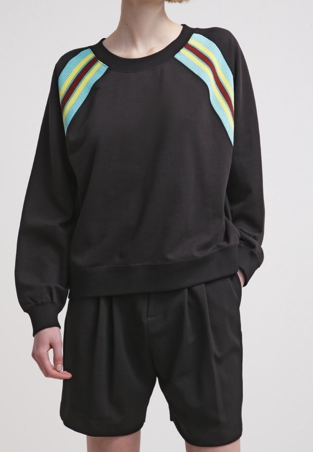 Personne portant un sweat-shirt noir avec des bandes bleues, jaunes et rouges sur les épaules, associée à un short noir ample, debout avec une main dans la poche.