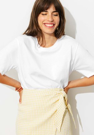 Jeune femme souriante, les yeux fermés, portant un t-shirt blanc et une jupe jaune à carreaux nouée à la taille, debout les mains sur les hanches.