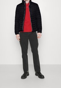 Sort pufferjakke med vandrette syninger, lynlås foran, rød polo-shirt under, grå bukser og sorte sko.