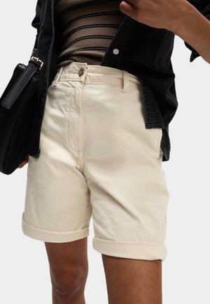 Person, die beige Shorts mit umgeschlagenem Saum, ein gestreiftes braunes Oberteil, eine schwarze Jacke trägt und eine schwarze Handtasche hält, vor einem einfarbigen Hintergrund.