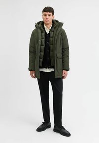 Olijfgroen pufferjack met rits en capuchon over een zwarte cardigan en een witte blouse, gecombineerd met zwarte broeken en schoenen.