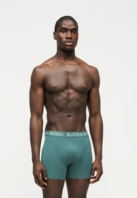 Αρσενικό μοντέλο με μυώδες σώμα που φοράει πετρόλ μπόξερ Björn Borg, στέκεται μπροστά σε απλό φωτεινό φόντο, κοιτάζοντας προς τα εμπρός.