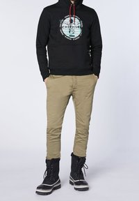 Svart hoodie med grafiskt tryck, kaki tapered byxor och svarta stövlar med vit kant och snörning. Tygernas texturer varierar mellan plagg.