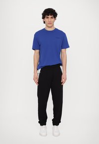 Μπλε κοντομάνικο μπλουζάκι με στρογγυλή λαιμόκοψη, συνδυασμένο με μαύρες φόρμες με κορδόνι και πλαϊνές τσέπες με φερμουάρ. Λευκά αθλητικά παπούτσια.