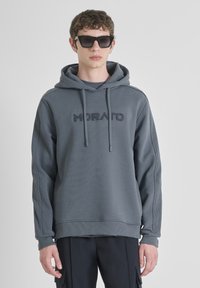 Felpa grigia con cappuccio, caratterizzata da un logo "MORATO" ricamato, cordini, polsi a costine e una vestibilità comoda, indossata con pantaloni neri e occhiali da sole.