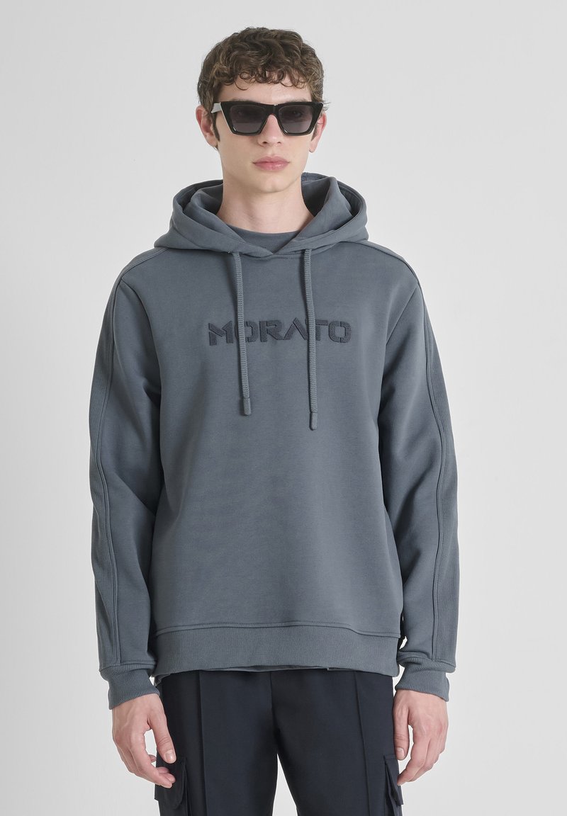 Felpa grigia con cappuccio, caratterizzata da un logo "MORATO" ricamato, cordini, polsi a costine e una vestibilità comoda, indossata con pantaloni neri e occhiali da sole.