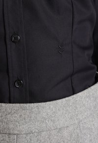 Camicia nera con bottoni e dettaglio ricamato a rosa; abbinata a un tessuto grigio testurizzato in vita, evidenziando materiali a contrasto.